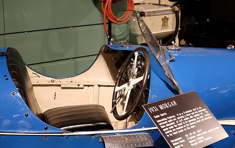 Close view of a blue three-wheeler’s cockpit with a four-spoke steering wheel and a black placard describing the car. Opens a larger view
