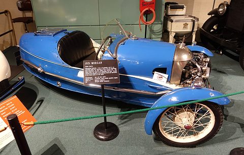 A bright blue three-wheeler with black seat and placard beside it, a green rope barrier in front. Opens a larger view