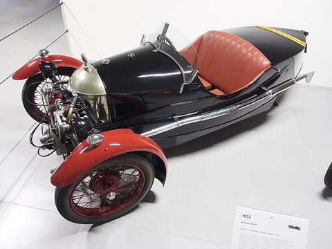 A black three-wheeler with red wings, red leather seats and an exposed V-twin on a white floor, a placard beside it. Opens a larger view
