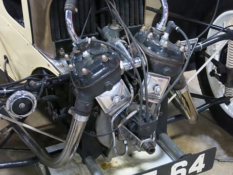 Close view of an air-cooled V-twin with chrome exhaust pipes, cables and a number plate below, cream wings either side. Opens a larger view