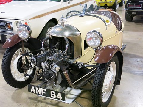 A cream three-wheeler with a polished cowl, winged mascot, brown wings and exposed V-twin, other cars behind. Opens a larger view