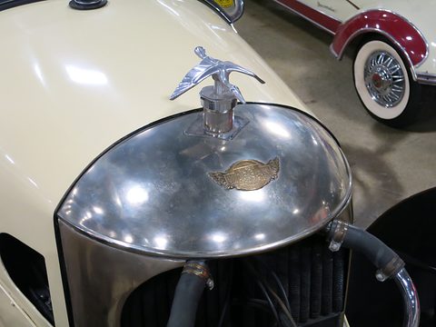 Close view of a polished domed cowl with a winged enamel badge and a flying mascot, cream bodywork around it. Opens a larger view