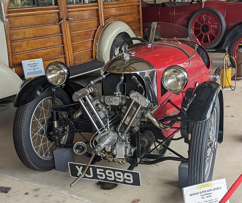 A red three-wheeler with a JAP V-twin, two large headlamps and black wings beside a wood-bodied estate car. Opens a larger view