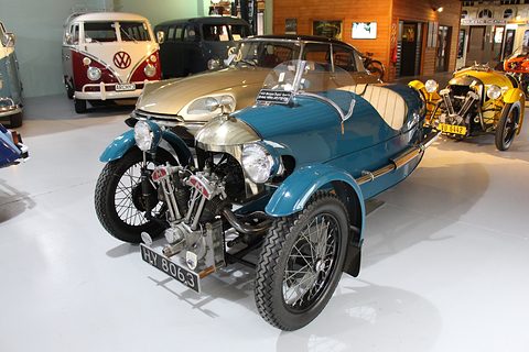 A teal three-wheeler with a Matchless V-twin and black wings on a white floor, a VW bus and other cars behind. Opens a larger view