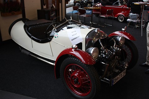 A white three-wheeler with maroon wings and wheels and an exposed V-twin on dark carpet. Opens a larger view