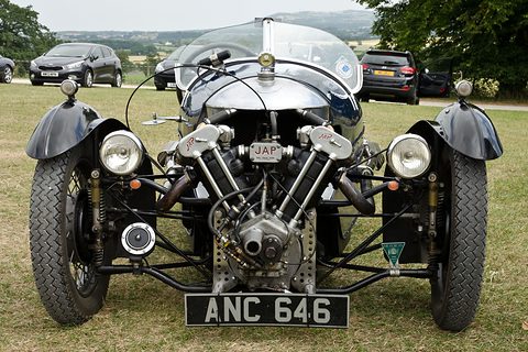 Front view of a three-wheeler with an exposed V-twin, two headlamps and black wings, modern cars behind. Opens a larger view
