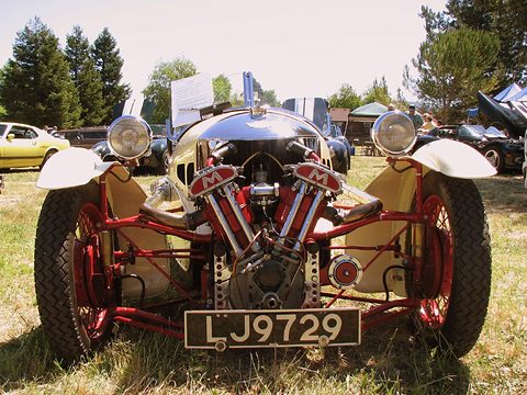Front view of a cream three-wheeler with red M-badged V-twin, red frame and two headlamps on grass. Opens a larger view