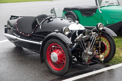 A black beetle-backed three-wheeler with red wire wheels and exposed V-twin on wet tarmac, a green car behind. Opens a larger view