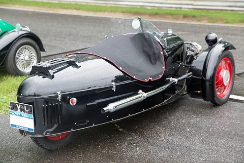 Rear quarter view of a black three-wheeler with a tonneau over the cockpit and a rally plate on the tail. Opens a larger view
