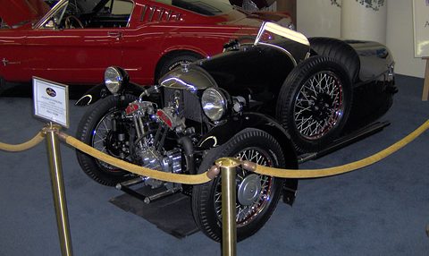 A black three-wheeler with an exposed V-twin and wire wheels on blue carpet behind brass posts and a rope. Opens a larger view