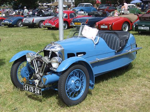 A light blue three-wheeler with exposed V-twin and black seat on grass, rows of classic sports cars behind. Opens a larger view