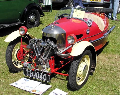 A red three-wheeler with cream wings, a JAP V-twin and badges on its front bar, parked on grass. Opens a larger view