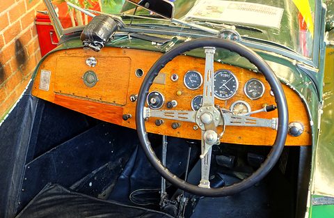 A varnished wooden dashboard with round dials behind a black four-spoke steering wheel. Opens a larger view