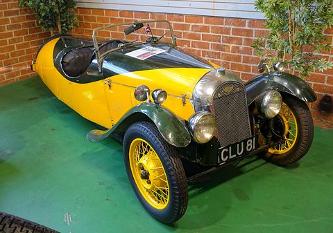 A yellow and dark green three-wheeler with a mesh grille, yellow wire wheels and a small Union flag, on green floor before a brick wall. Opens a larger view