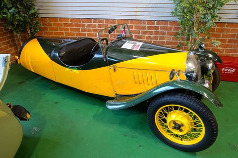 Side view of a yellow three-wheeler with dark green wings, louvred bonnet and yellow wire wheels before a brick wall. Opens a larger view