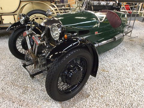 A dark green beetle-backed three-wheeler with black wings and an exposed V-twin on a white gravel display. Opens a larger view