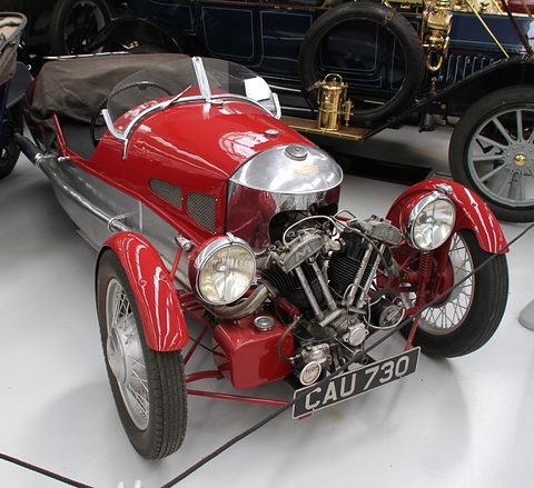 A red and silver three-wheeler with an exposed V-twin, two headlamps and a black hood on its tail, on a white floor. Opens a larger view