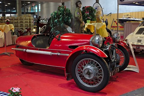 A red three-wheeler with wire wheels and chrome side pipes on red carpet, mannequins and plants behind. Opens a larger view