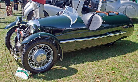 A glossy dark green three-wheeler with a barrel-shaped tail, spare wheel and wire wheels on grass. Opens a larger view