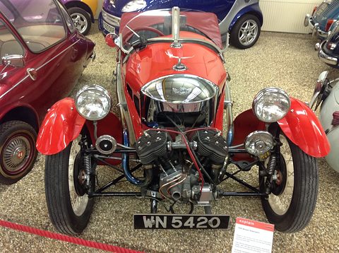 Front view of a red three-wheeler with a polished cowl, V-twin and two headlamps on gravel among microcars. Opens a larger view