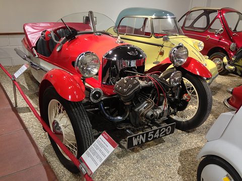 A red and cream three-wheeler with exposed V-twin on gravel behind a rope, bubble cars beside it. Opens a larger view