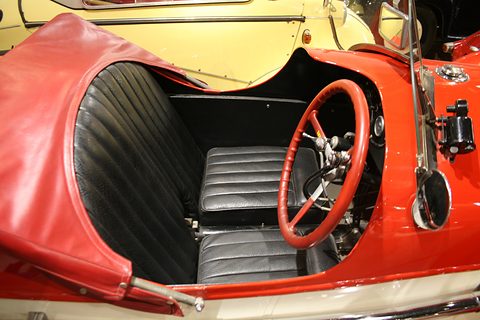 Black pleated leather seats and a red steering wheel inside a red three-wheeler with its hood folded. Opens a larger view