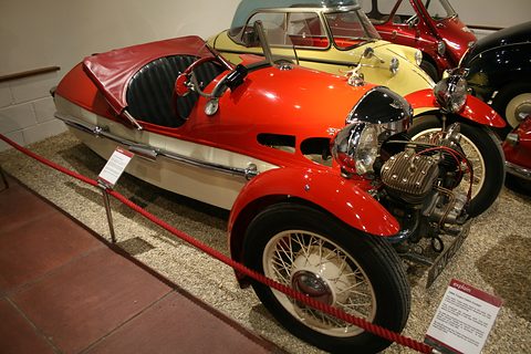 A red and cream three-wheeler with folded hood and cream wire wheels behind a red rope, a placard in front. Opens a larger view
