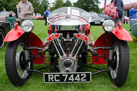 Front view of a red three-wheeler with a chrome cowl, exposed V-twin and two headlamps on grass. Opens a larger view