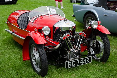 A red three-wheeler with black seats, chrome cowl and exposed V-twin on grass beside a pale blue car. Opens a larger view
