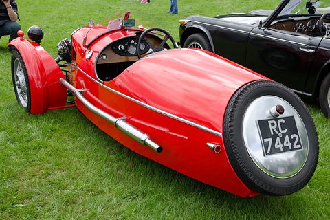 Rear quarter view of a red three-wheeler with a rounded tail carrying a chrome-covered spare wheel and plate. Opens a larger view