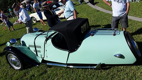 Side view of a pale green three-wheeler with a long louvred bonnet, swept wings and black hood, people on grass behind. Opens a larger view