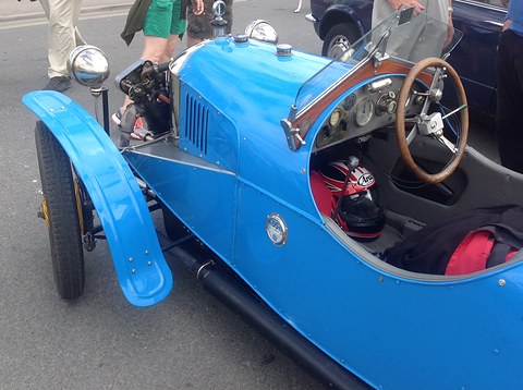 A bright blue three-wheeler seen from above the cockpit: wooden steering wheel, dashboard gauges and a helmet on the seat. Opens a larger view