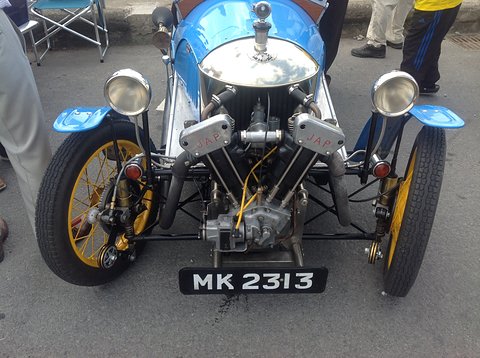Front view of a bright blue three-wheeler with a polished cowl, two headlamps, a V-twin and yellow wheels. Opens a larger view