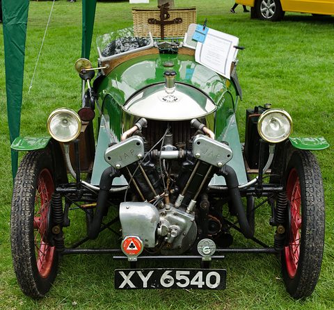 Front view of a green three-wheeler with a JAP V-twin, two brass headlamps, red wheels and a picnic hamper on the tail. Opens a larger view
