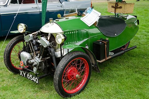A green three-wheeler with an exposed JAP V-twin, red wire wheels and a hamper on the tail, parked on grass. Opens a larger view