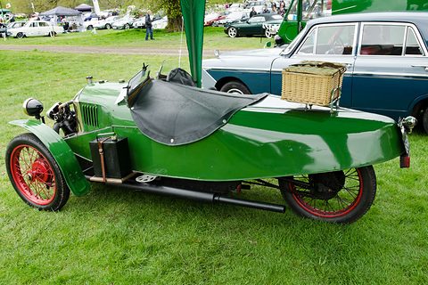 Side view of a green three-wheeler with red wheels, a black tonneau and a wicker hamper on the tail, a blue saloon behind. Opens a larger view