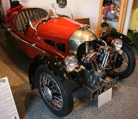 A red three-wheeler with black wings, an exposed V-twin, two headlamps and black seats, a placard on a stand. Opens a larger view