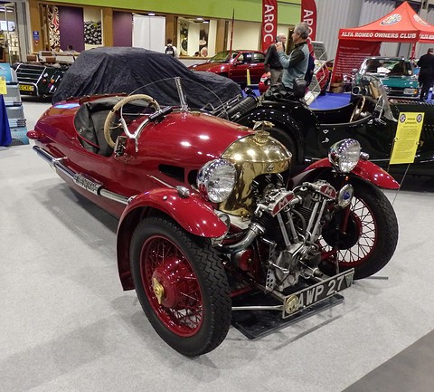 A maroon three-wheeler with a brass cowl, exposed V-twin and red wire wheels on grey carpet, visitors behind. Opens a larger view