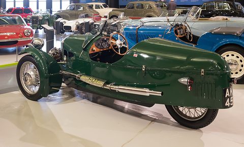 A dark green three-wheeler from behind and the side, wooden dashboard visible, other cars in a bright hall. Opens a larger view