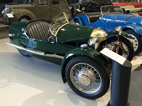 Side view of a dark green beetle-backed three-wheeler with black seats, a blue racing car behind. Opens a larger view