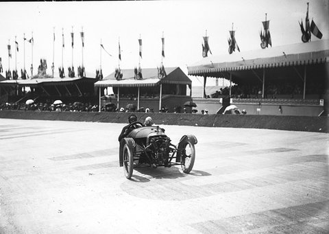 A three-wheeled racing cyclecar with an exposed V-twin at its nose and two crew, on a concrete track before decorated stands.