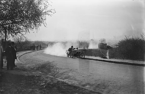 A low three-wheeler with two crew sliding round a curving road in a cloud of dust, onlookers in coats under a tree. Opens a larger view