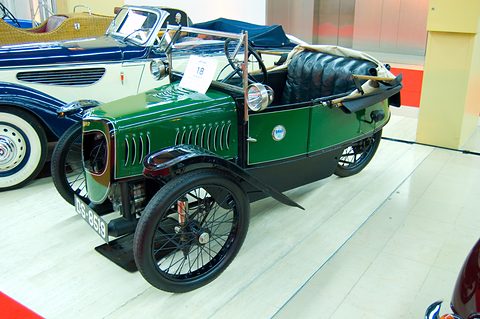 A green three-wheeler with a louvred bonnet, black wings and black seats on a white floor beside a blue saloon. Opens a larger view
