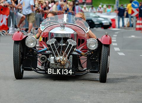 Front view of a red three-wheeler with a big JAP V-twin and two headlamps, two women aboard and spectators behind. Opens a larger view
