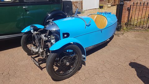 A bright blue three-wheeler with an exposed V-twin and black wings on a brick-paved drive, a dark green car behind. Opens a larger view