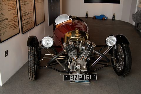 Front view of a maroon three-wheeler with an exposed V-twin, two headlamps and a brass cowl in a white room. Opens a larger view