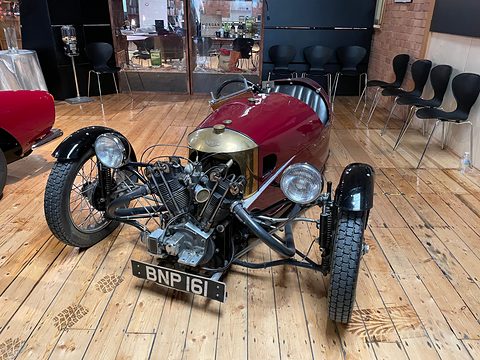 A red three-wheeler with an exposed V-twin, brass cowl and two headlamps on a wooden floor before black chairs. Opens a larger view