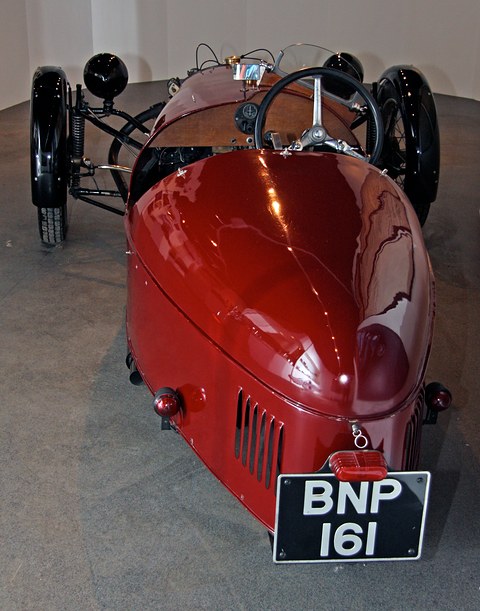 Rear view of a maroon three-wheeler with a rounded tail, louvres and a number plate, steering wheel visible beyond. Opens a larger view