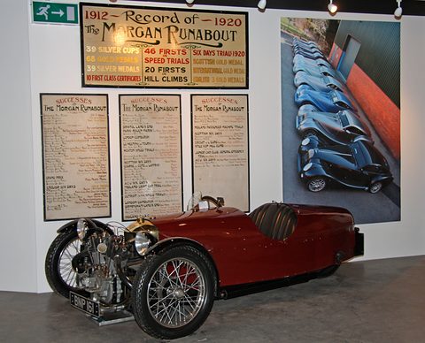 A maroon three-wheeler on a floor before signboards listing silver cups and speed trials, and a poster of blue Morgans. Opens a larger view