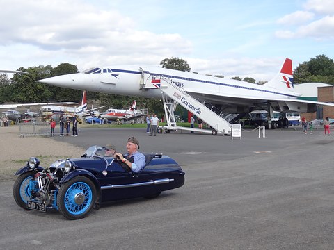 A dark blue three-wheeler with two men aboard on tarmac, Concorde parked behind under a cloudy sky. Opens a larger view